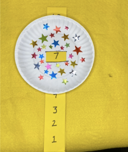 White paper plate decorated with star stickers with a paper strip with numbers ten to one visible through a hole in the middle on a yellow background